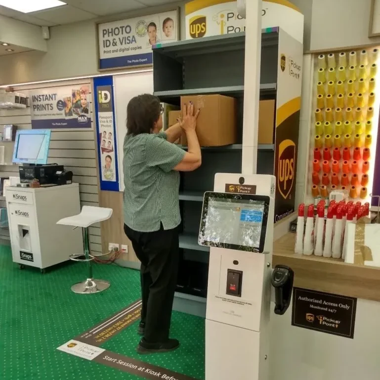 Person placing package in automated UPS Access Point locker at retail store with tablet interface in foreground.