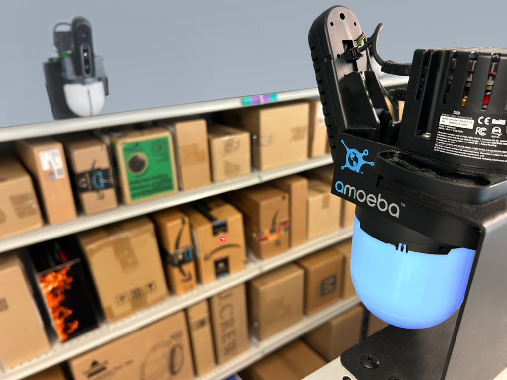 Amoeba robotic arm with blue illuminated base positioned near shelving units filled with cardboard boxes in a warehouse or storage setting.