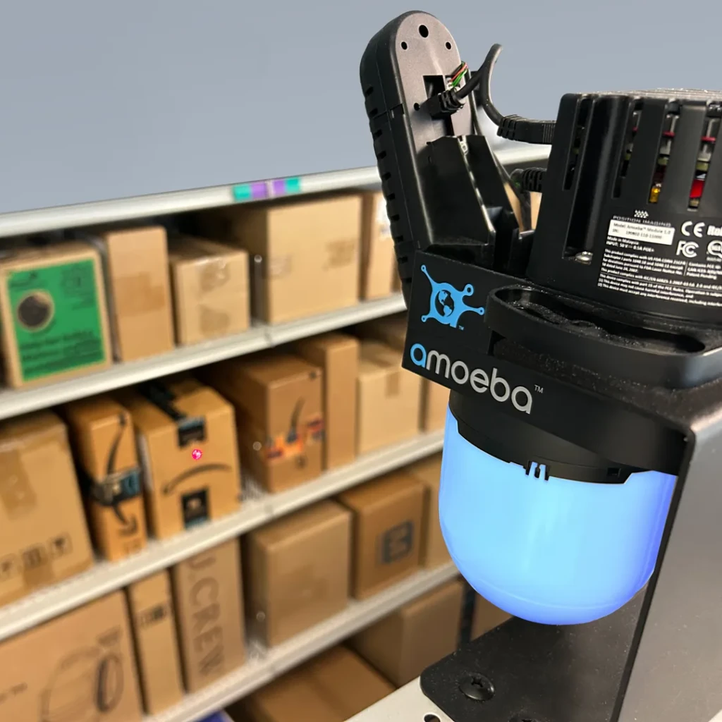 Amoeba robotic arm with blue illuminated base positioned near shelving units filled with cardboard boxes in a warehouse or storage setting.