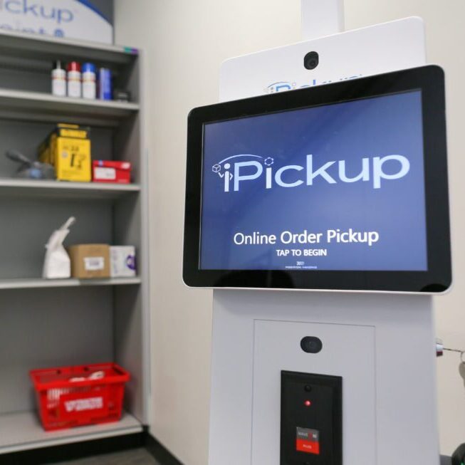 iPickup kiosk displaying "Online Order Pickup" screen next to shelving unit with retail products