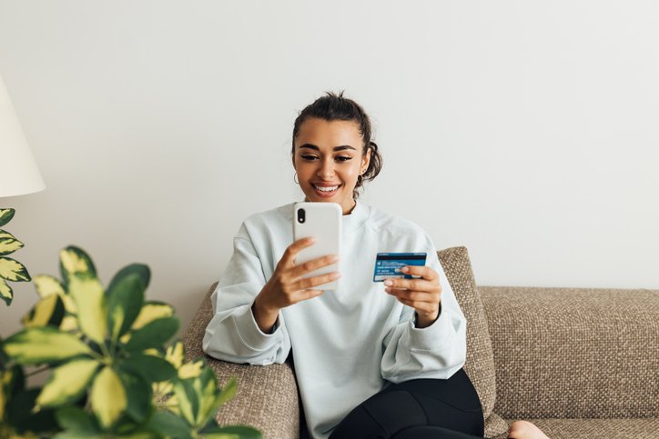 Smiling woman on a couch holding a credit card while shopping on her phone